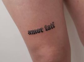 Маленьке тату "amor fati" на нозі, виконане чорним шрифтом у готичному стилі, символізує прийняття долі та життєвий філософський підхід. Тату "amor fati" на ноге, стиль чорнильного шрифту.