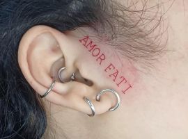 Модель з татуюванням "Amor Fati" на вусі, доповнена кількома ювелірними виробами, підкреслює індивідуальність і глибокий зміст. Тату "Amor Fati" на вусі з червоним шрифтом, символізує прийняття долі та життєвий філософський підх.