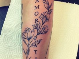 Підпис до зображення: Татуювання "Amor Fati" з квітами на передпліччі, символ прийняття долі та життєвої філософії. Татуювання "Amor Fati" з квітами на руці, стильне та значуще.