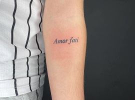 Маленьке тату "Amor fati" на руці, символ прийняття долі та філософії стоїцизму, створене у тату-салоні Альянс. Тату "Amor fati" на передпліччі, чорний шрифт, мінімалістичний дизайн.