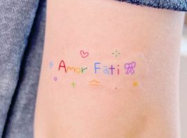 Маленьке яскраве тату "Amor Fati" на руці, що символізує прийняття своєї долі та життєвий філософський підхід. Барвистий тату "Amor Fati" на руці, символ прийняття долі.