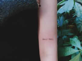 Малюнок "Amor Fati" на руці, символ прийняття долі та філософії життя, у природному оточенні. Тату "Amor Fati" на руці біля листя рослини.